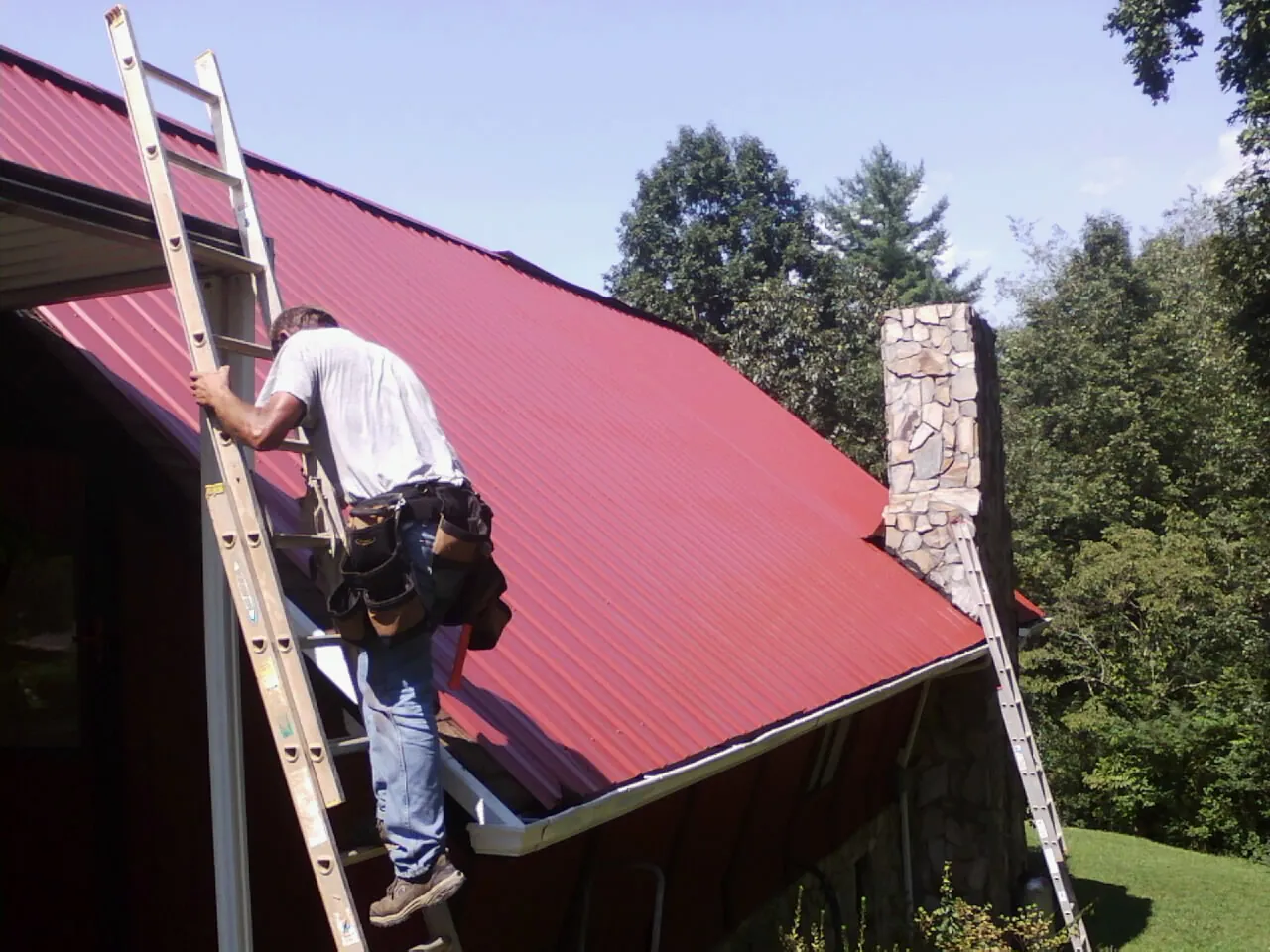 Roofer on a ladder beside a red metal roof and stone chimney — Soffit & Fascia Repair by Roofing Brothers in Prattville